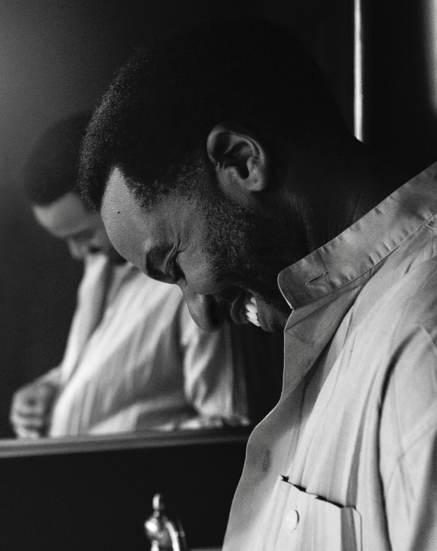 A black and white photo of actor André Holland’s left side profile as he smiles while buttoning up his shirt in front of a mirror. He is a mid-40s Black man with a small Afro and smooth skin.