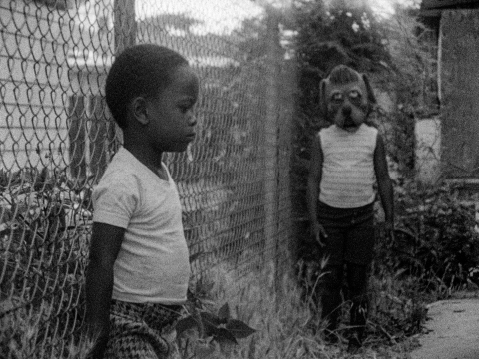 In Killer of Sheep, two young Black children against a fence. One of them has their back facing the fence, while the other is standing a few feet away wearing a caricatured dog mask.