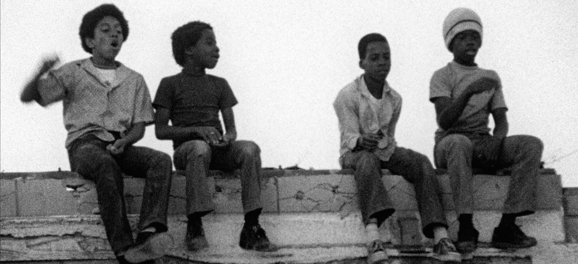 A still from the Killer of Sheep. Four young Black boys sit on a brick wall. One appears to be cheering, and another faces away from the camera. The other two are looking down at something.