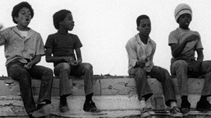 A still from the Killer of Sheep. Four young Black boys sit on a brick wall. One appears to be cheering, and another faces away from the camera. The other two are looking down at something.