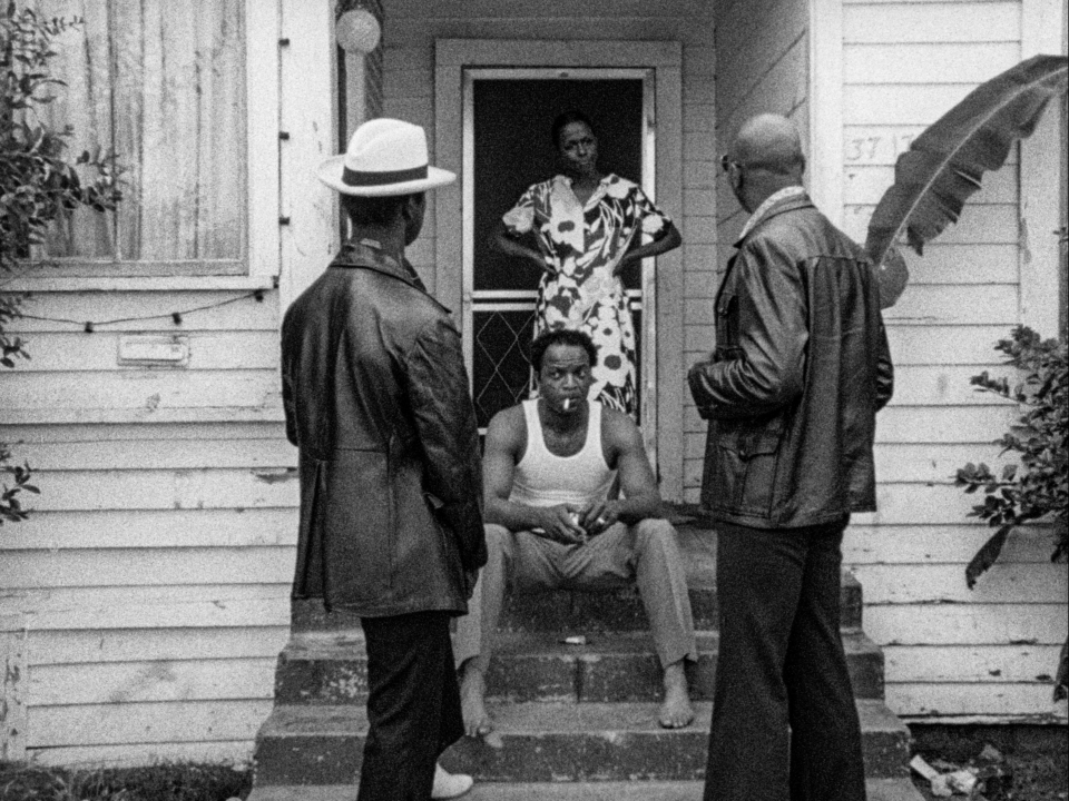 A still from the film Killer of Sheep (1978) by Charles Burnett. Two men stand facing the doorway of a house. A man is sitting on the steps, and a woman stands behind him with her hands on her hips. All of them are Black.