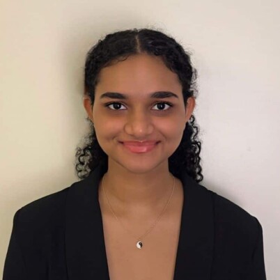 A headshot of Ritikaa. They are a women of color, they have on a black blazer and a silver necklace. They are smiling at the camera.