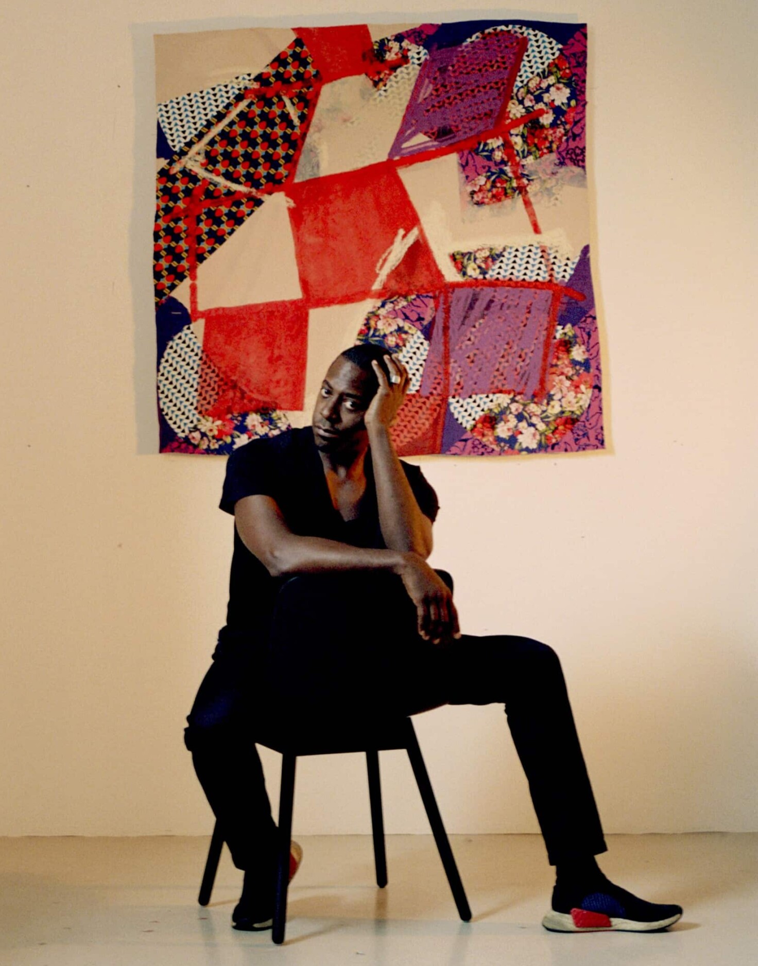 Sanford Biggers sits backwards on a chair while wearing all black with a quilted artwork on the wall behind him. The work is a patchwork of differently patterned fabrics and red quadrilaterals painted on top.