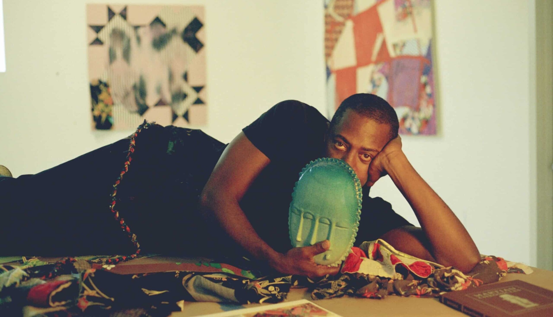 Sanford Biggers lying down on his side with one hand holding his head up, and the other holding an art piece in the shape of a sculpted teal mask that covers the bottom half of the face.