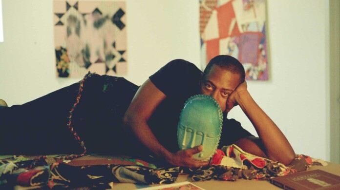 Sanford Biggers lying down on his side with one hand holding his head up, and the other holding an art piece in the shape of a sculpted teal mask that covers the bottom half of the face.