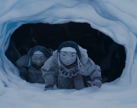 An animated still from The Shaman's Apprentice shows two Indigenous people crawling through a snowy cave. They are wearing large cloaks and have face paint. They have determined expressions.