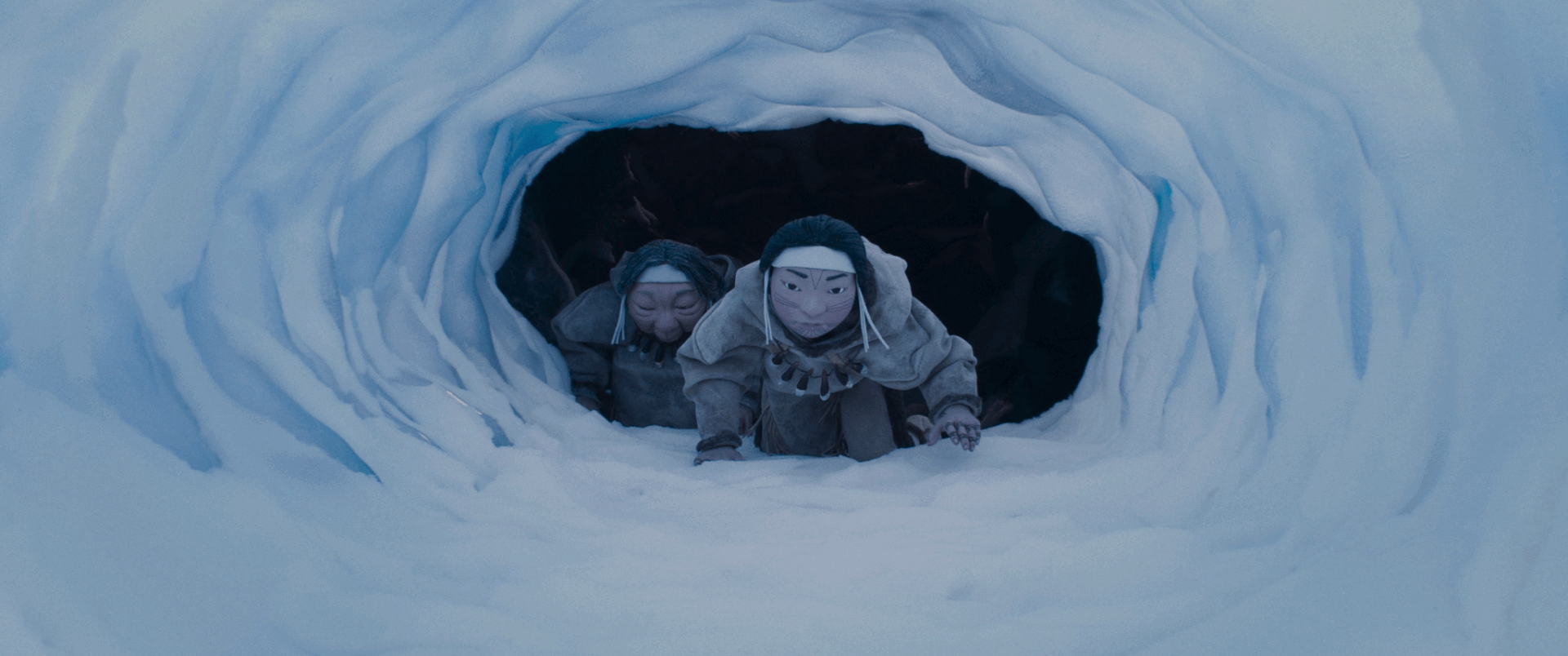 An animated still from The Shaman's Apprentice shows two Indigenous people crawling through a snowy cave. They are wearing large cloaks and have face paint. They have determined expressions.