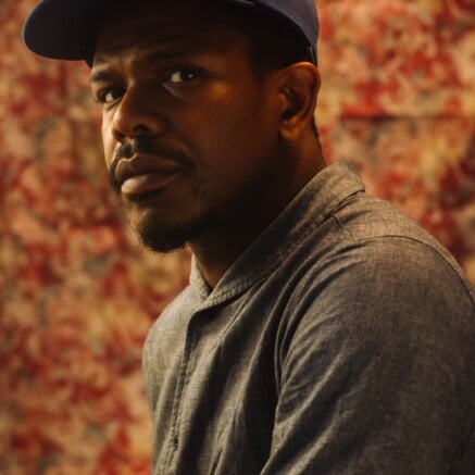 A portrait of Lendl Tellington. He is a Black man. He is wearing a navy blue cap and a gray button-down shirt. He is looking at the camera with a contemplative expression.