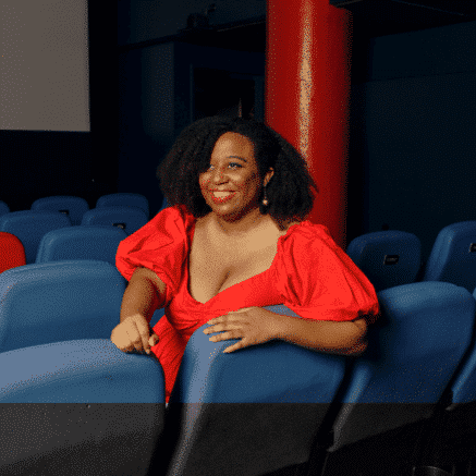 A photo of Maya S. Cade. She is a Black women. She is sitting in a movie theater. She is wearing a bright red dress. She has curly, long black hair.