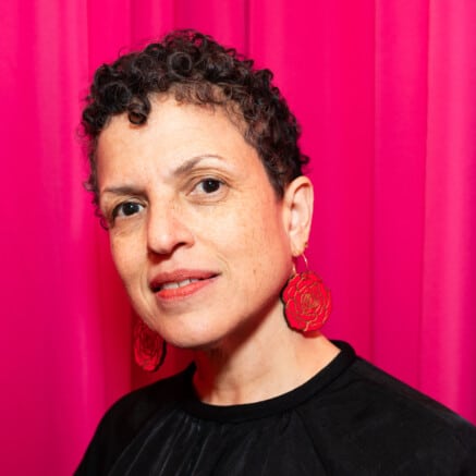 A headshot of Michèle Stephenson. She has short curly hair and is wearing red earrings. She is standing in front of a pink backdrop.