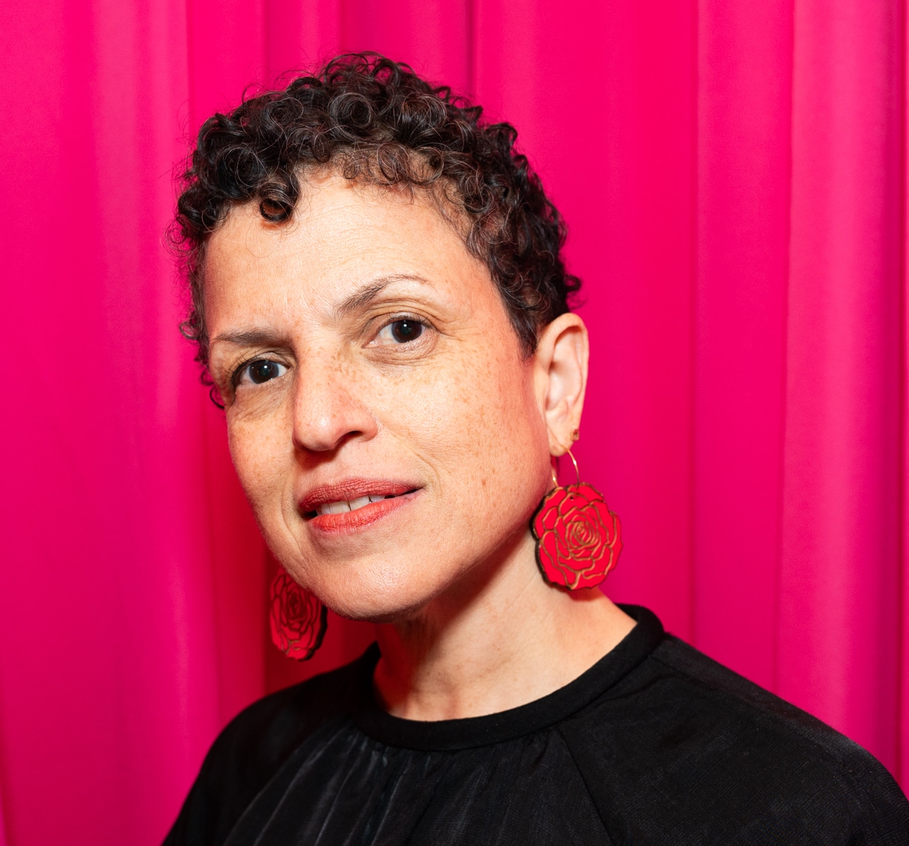A headshot of Michèle Stephenson. She has short curly hair and is wearing red earrings. She is standing in front of a pink backdrop.