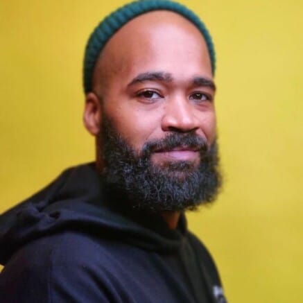 A headshot of Rashid Zakat. He is a Black man, he is wearing a green beanie and a black hoodie. He is looking at the camera with a slight smile.