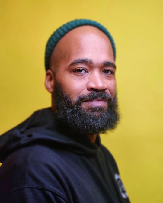 A headshot of Rashid Zakat. He is a Black man, he is wearing a green beanie and a black hoodie. He is looking at the camera with a slight smile.