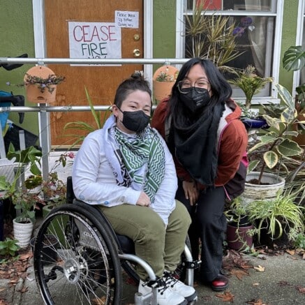 A photo of two people posing together outside of what appears to be a row home. One person is in a wheelchair and the other is standing beside them, both have on black KN95 face masks. There are signs on the door that read "Ceasefire Now" and "Please mask up before coming in."