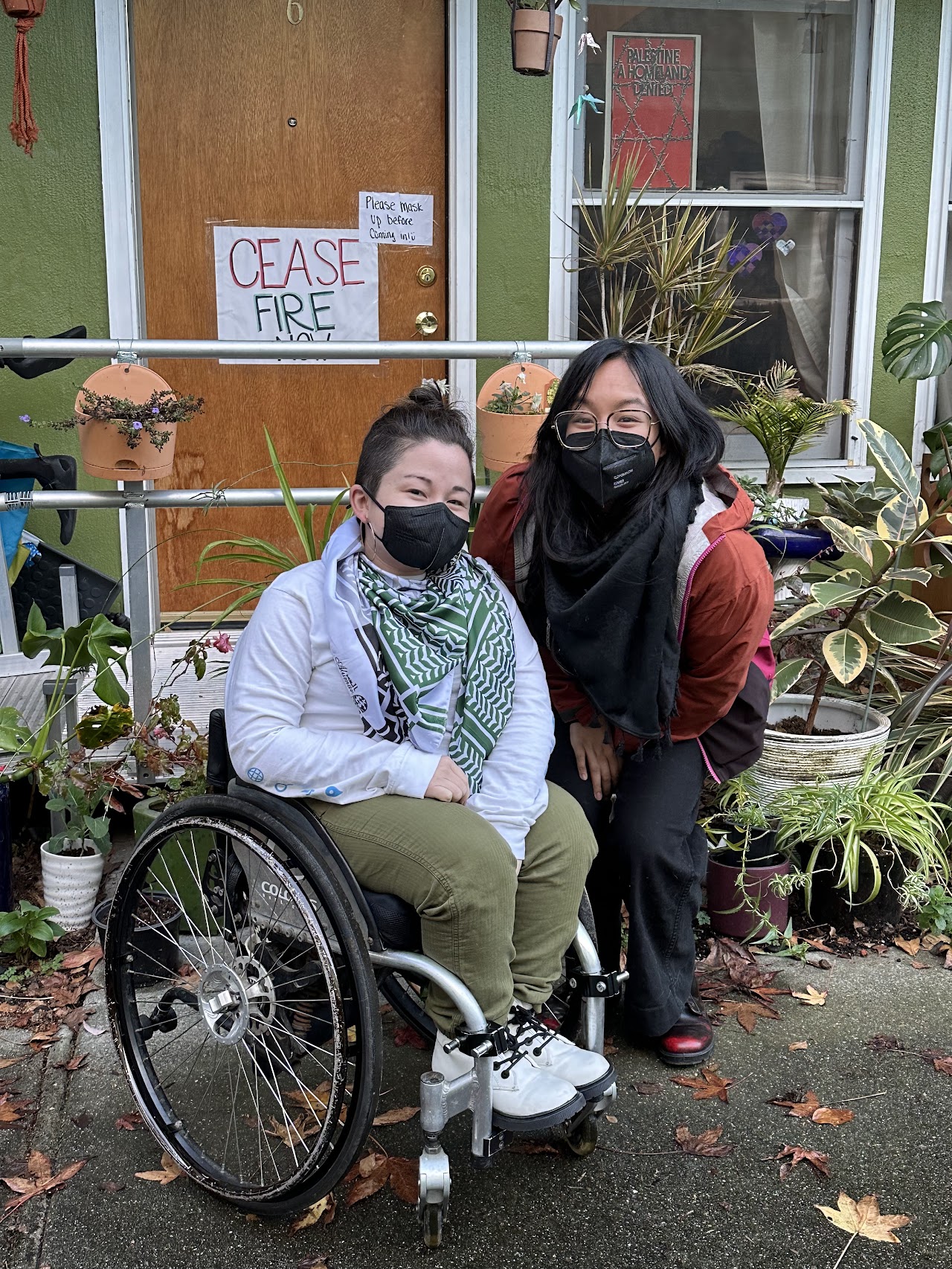 A photo of two people posing together outside of what appears to be a row home. One person is in a wheelchair and the other is standing beside them, both have on black KN95 face masks. There are signs on the door that read "Ceasefire Now" and "Please mask up before coming in."
