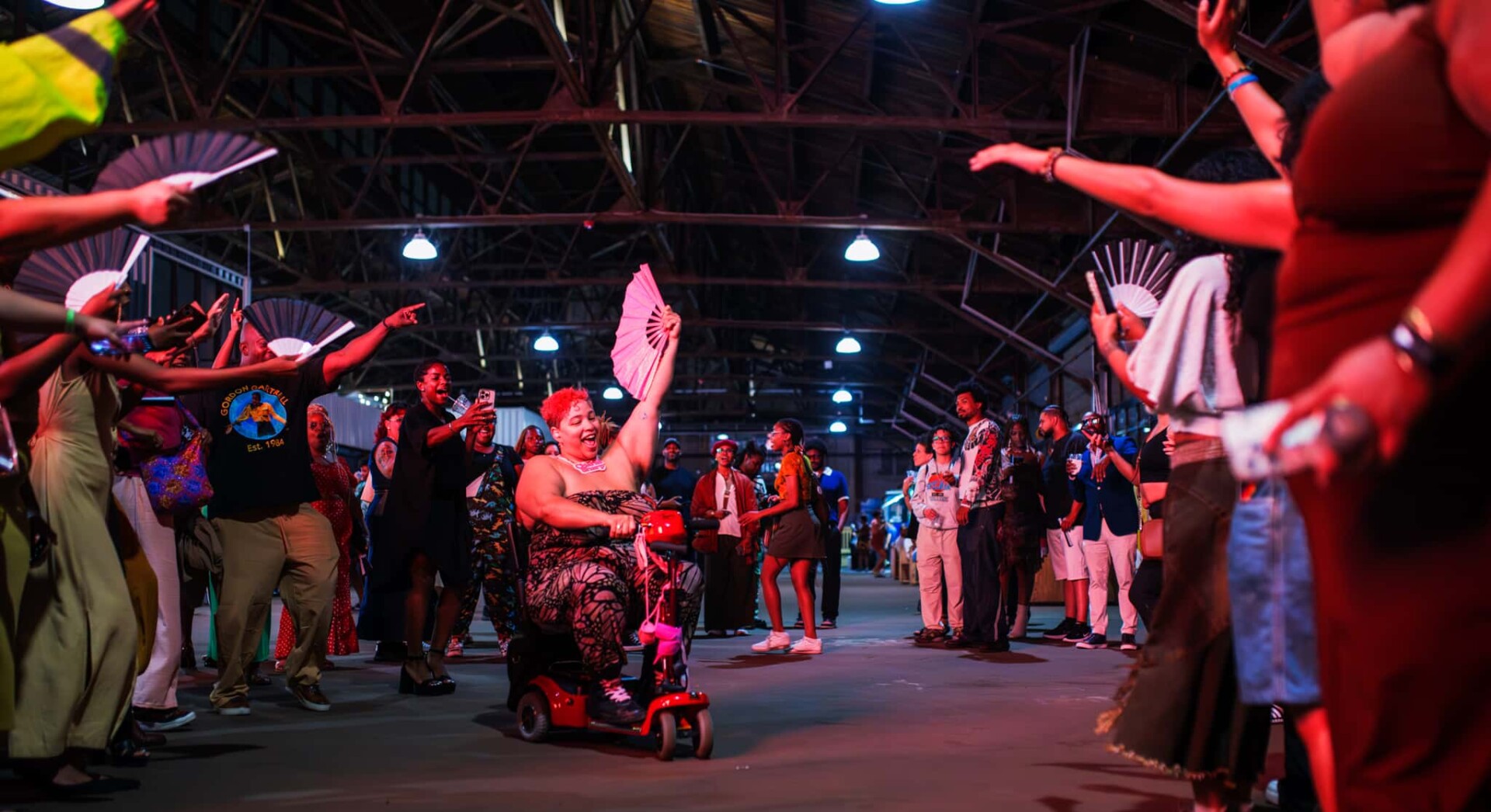 A photo from the 2025 BlackStar Film Festival opening party. A spirit tunnel has opened on the dance floor, a person in a wheelchair is going through it, they are holding a paper fan in the air. Those on the sides have paper fans as well, raised in celebration.