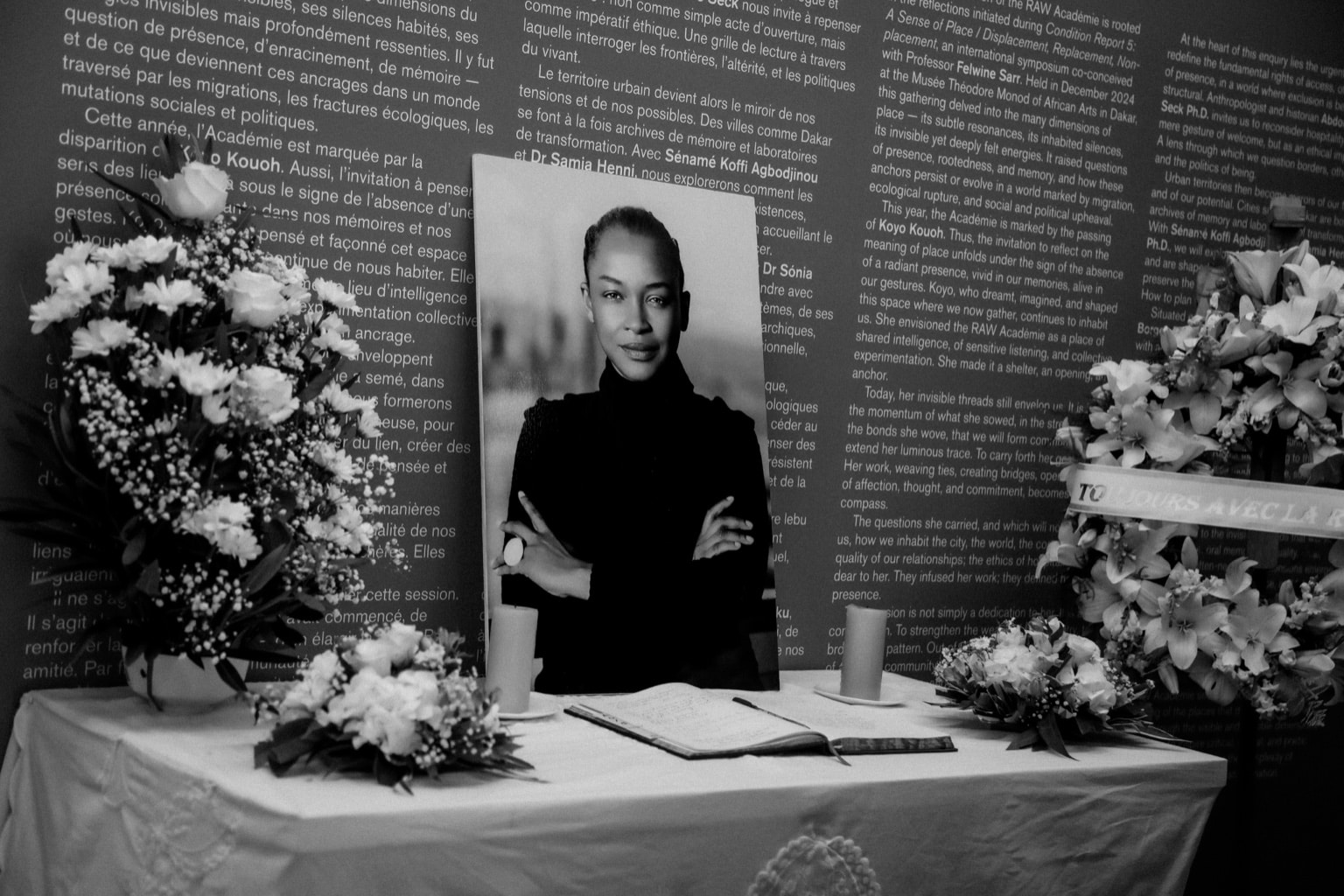 A black and white photo of a large photo of Koyo Kouoh leans against a wall in front of a guest book, surrounded by flowers at her memorial.