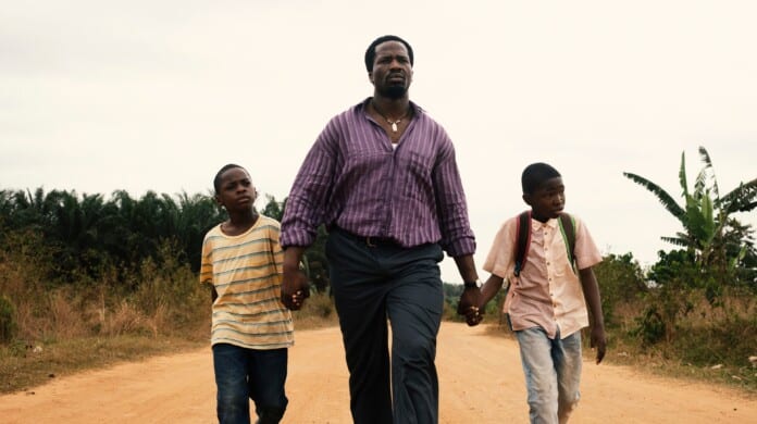A father is walking with his two sons across a deserted sandy road. The father stands in the middle, holding both of his sons hands as they walk together.