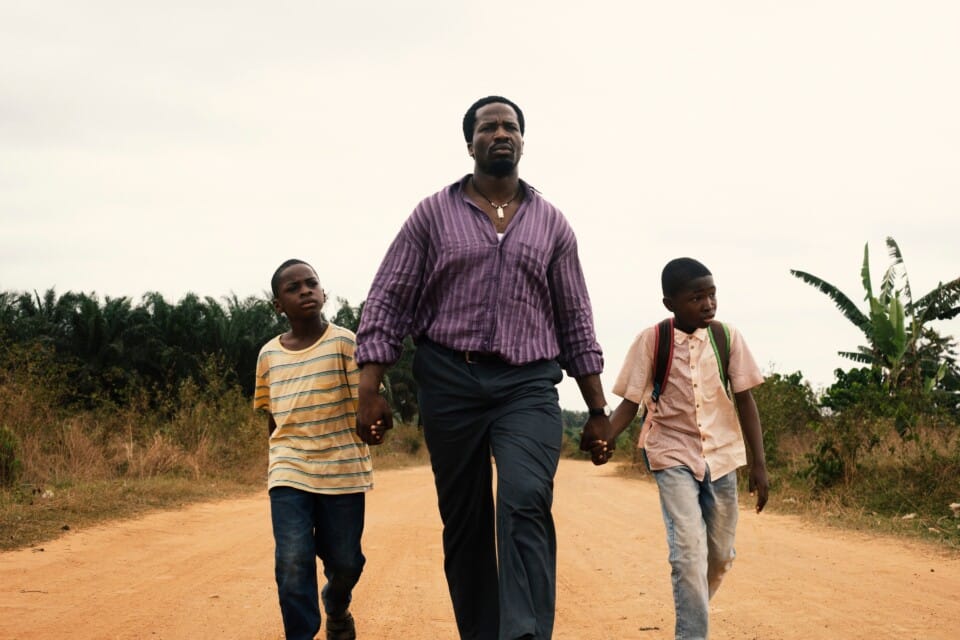 A father is walking with his two sons across a deserted sandy road. The father stands in the middle, holding both of his sons hands as they walk together.