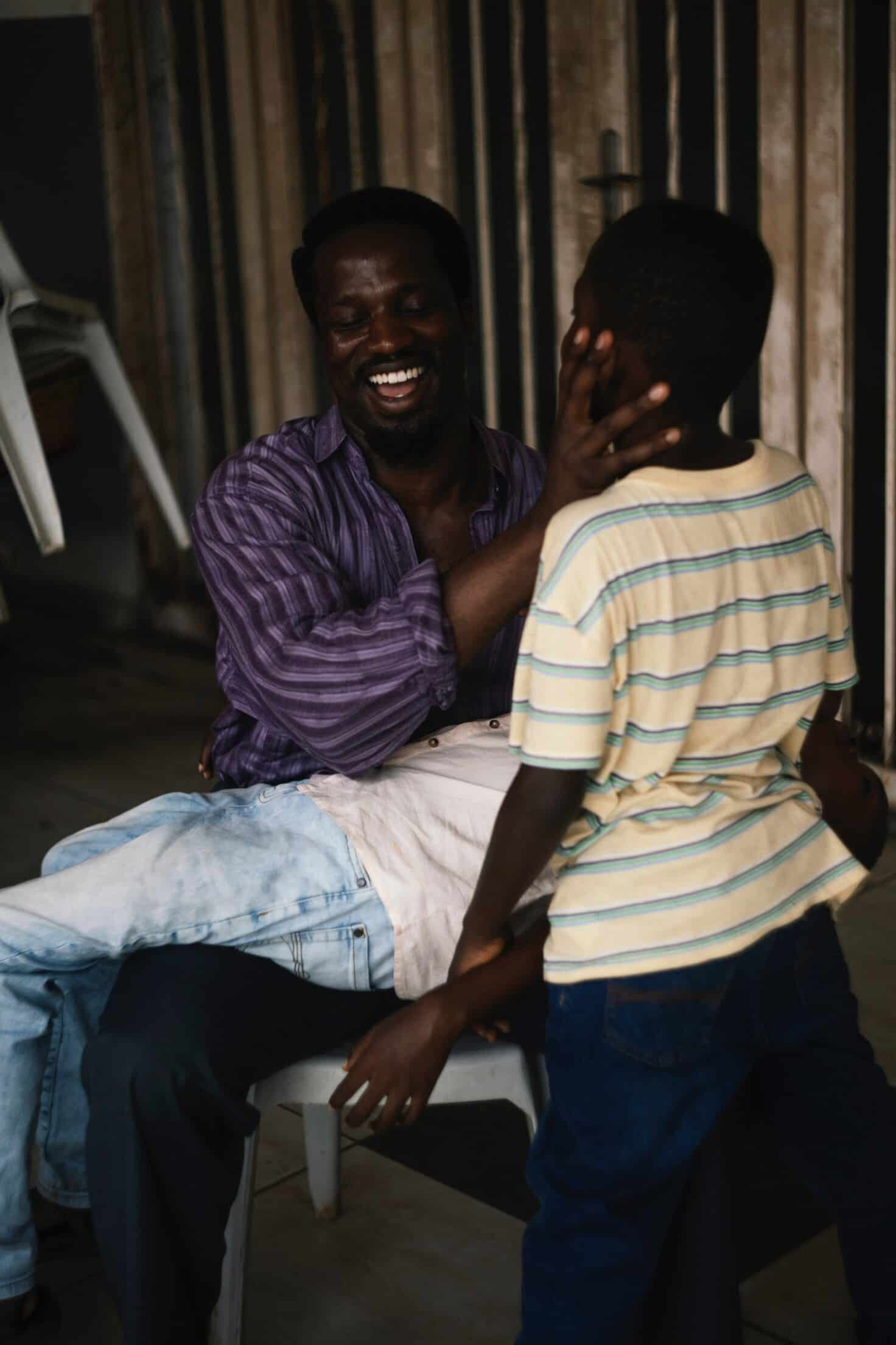 The dad is sitting in a chair, smiling, with one son lying on his lap and the other standing in front of him.