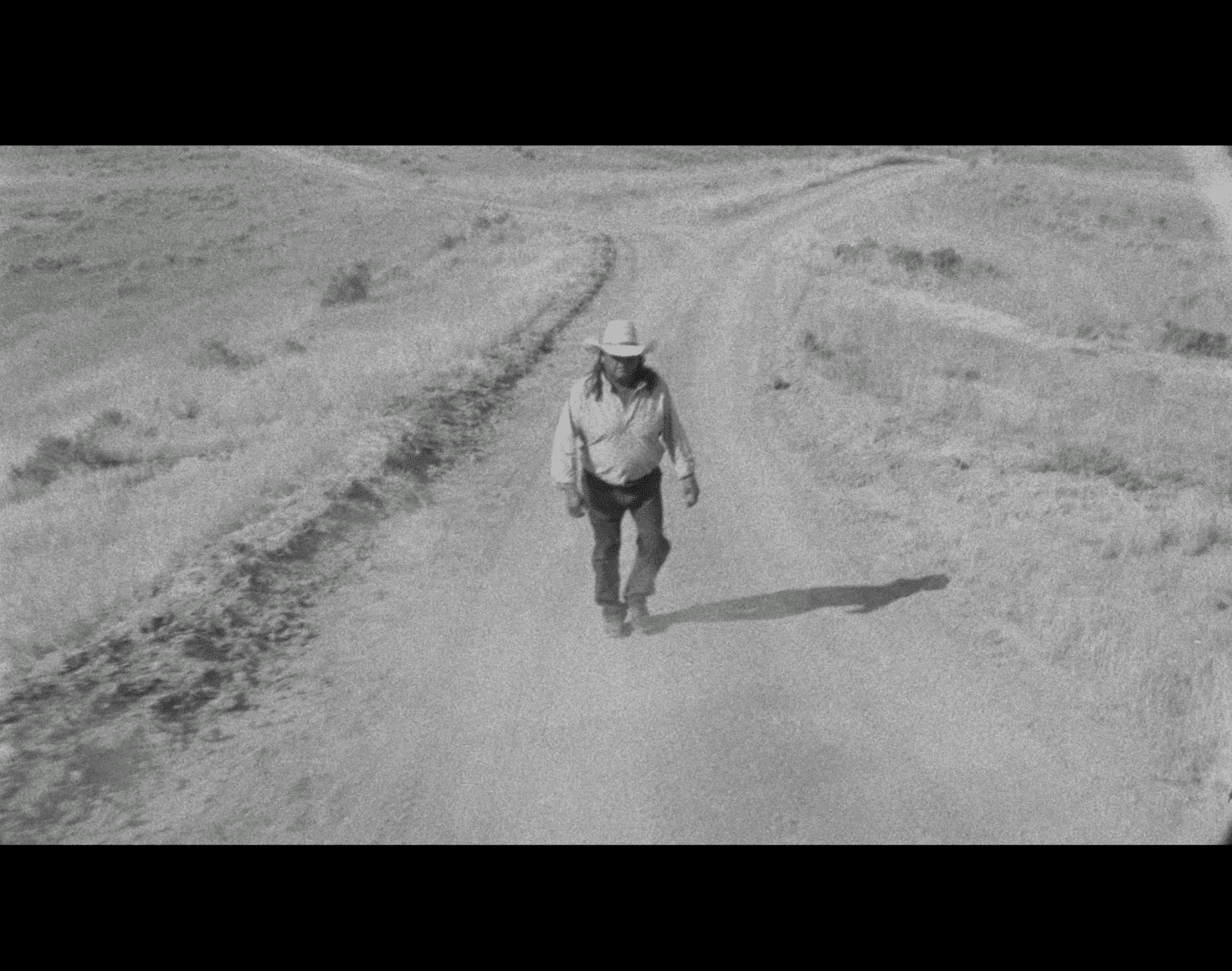 A black-and-white still from The Day the Sun Died shows a man walking down a dirt road. He's just passed a point where two roads have merged into one.