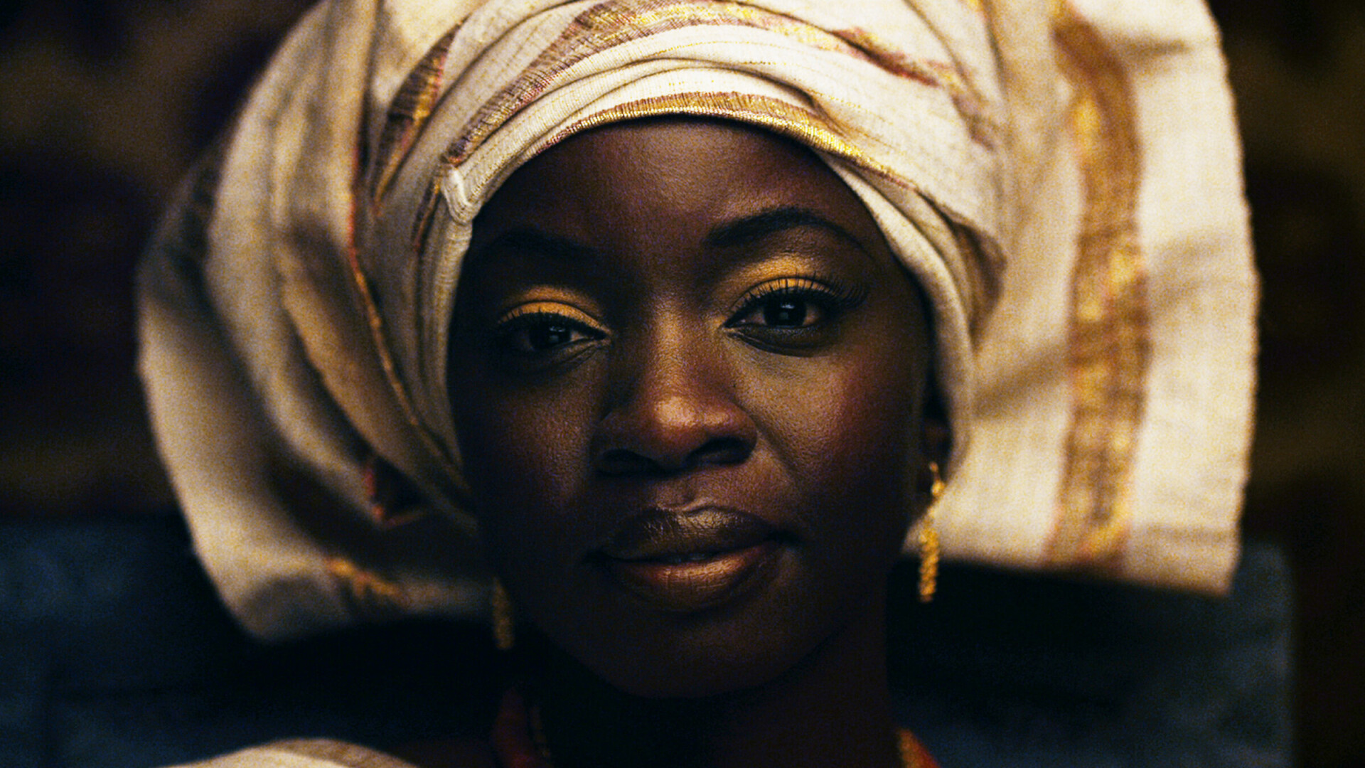 A film still from Mother of George. It is a close-up shot of the face of an African woman. She has gold eye shadow, earrings, and a gold-and-tan head wrap. She is looking directly at the camera with a soft but striking gaze.