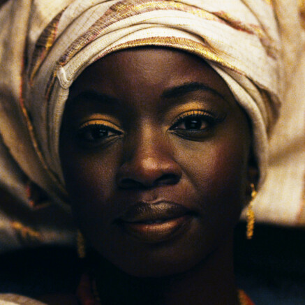 A film still from Mother of George. It is a close-up shot of the face of an African woman. She has gold eye shadow, earrings, and a gold-and-tan head wrap. She is looking directly at the camera with a soft but striking gaze.