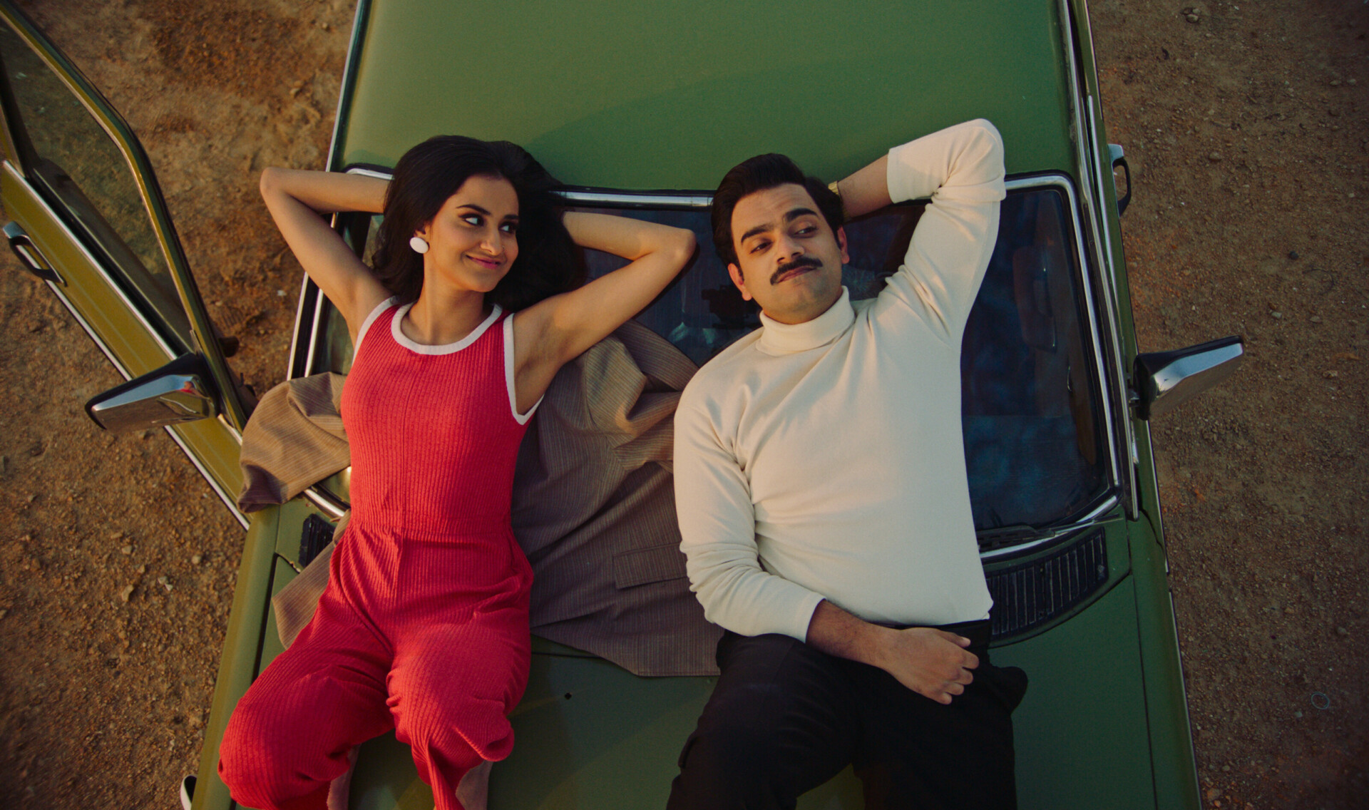 A still from The Queen of My Dreams shows two young people laying on the hood of an old car. The women is looking at the man with a soft smile. The man is looking up at the sky above him.