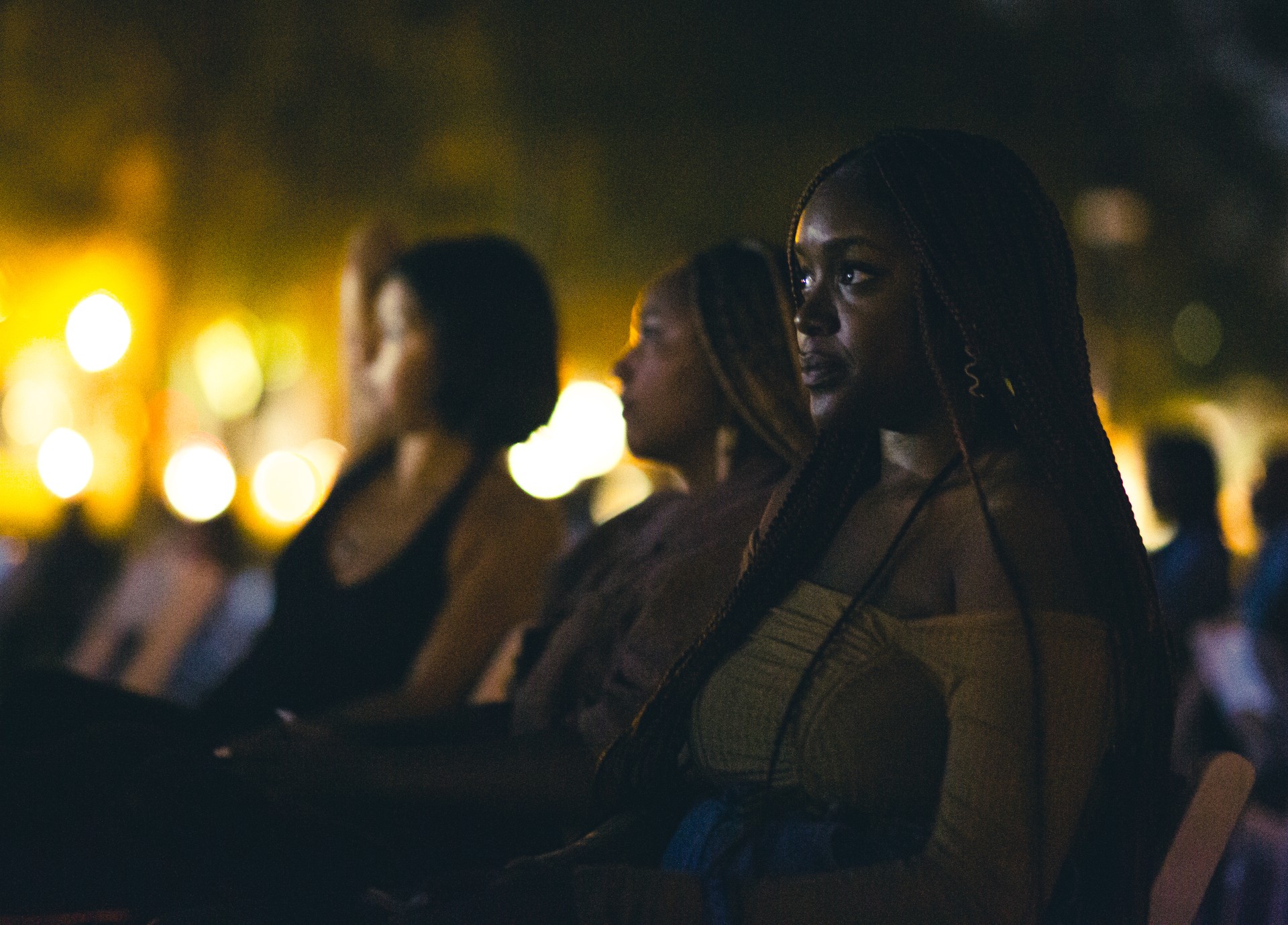A nightime photo of the audience during an outdoor film screening focuses on three people, the one in the foreground in focus, who is a Black person with long hair.
