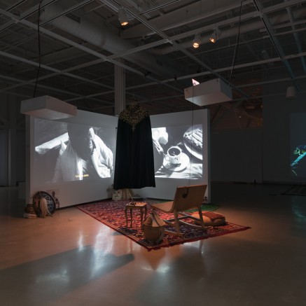 A photo of a gallery exhibition featuring multiple projected videos on white screens, plus a carpeted viewing area in the middle of an otherwise dark room.