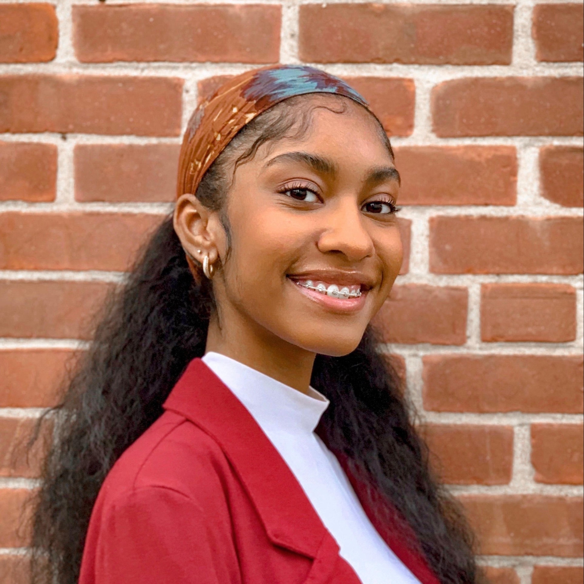 Cienna Benn smiles at the camera in front of a brown brick wall. She wears a red blazer over a white shirt.
