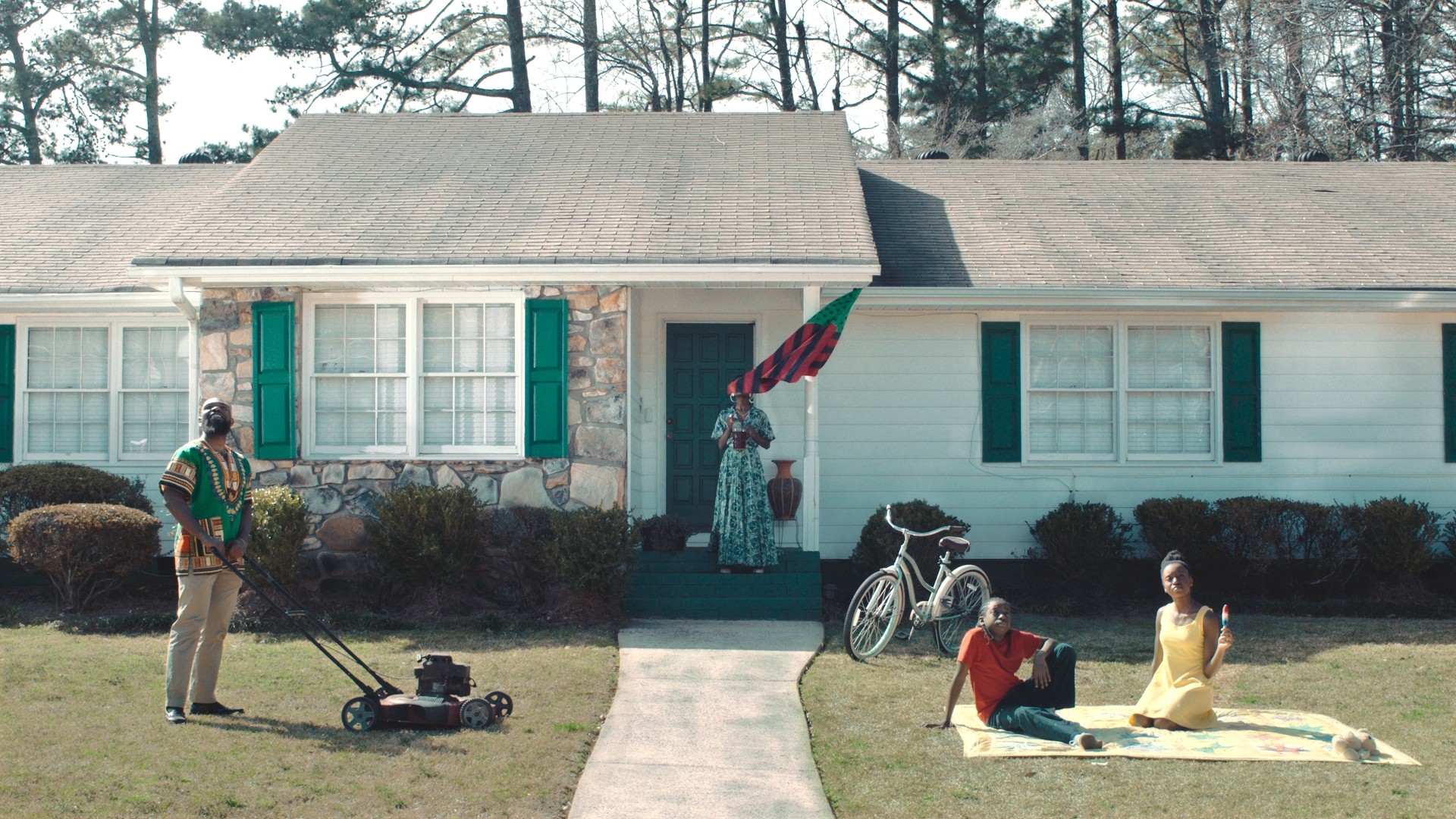Still from Eyes on the Prize: Hallowed Ground shows a Black family in a deceptively idyllic scene — two children having a picnic on one side of the lawn as a parent mows the other side. On the steps of the home stands the other parent, perhaps the mother, but their face is obscured by a an American flag represented in the colors of red, black, and green. The three people whose faces are visible are all looking up at something in the sky.