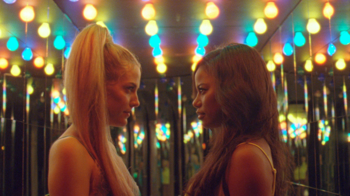Still from the film "Zola" shows the side profiles of two women as they face each other. Behind them, rows of rainbow light bulbs line the ceiling.