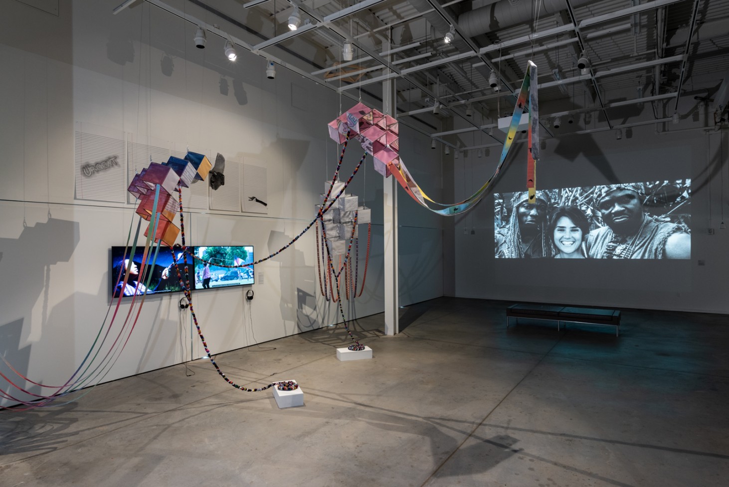 A photo of the Assemblage exhibit displays tv screens on the walls and colorful items hanging from the rafters inside a sparse, warehouse like space.