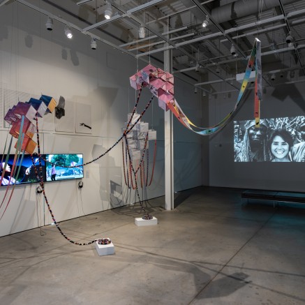 A photo of the Assemblage exhibit displays tv screens on the walls and colorful items hanging from the rafters inside a sparse, warehouse like space.