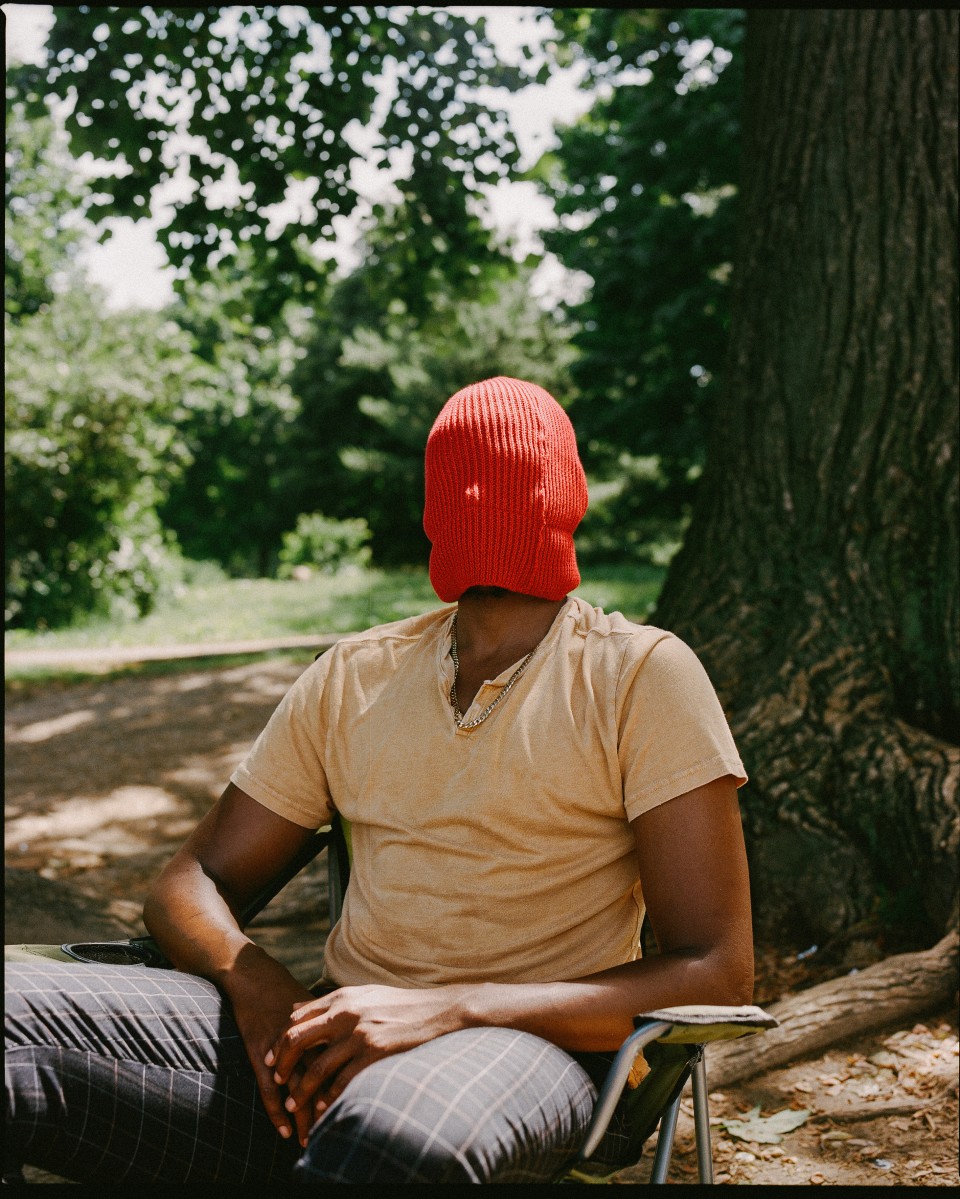 A Black person sits in a chair outside, in front of a tree, their face covered by an orange/green beanie pulled down to their neck.