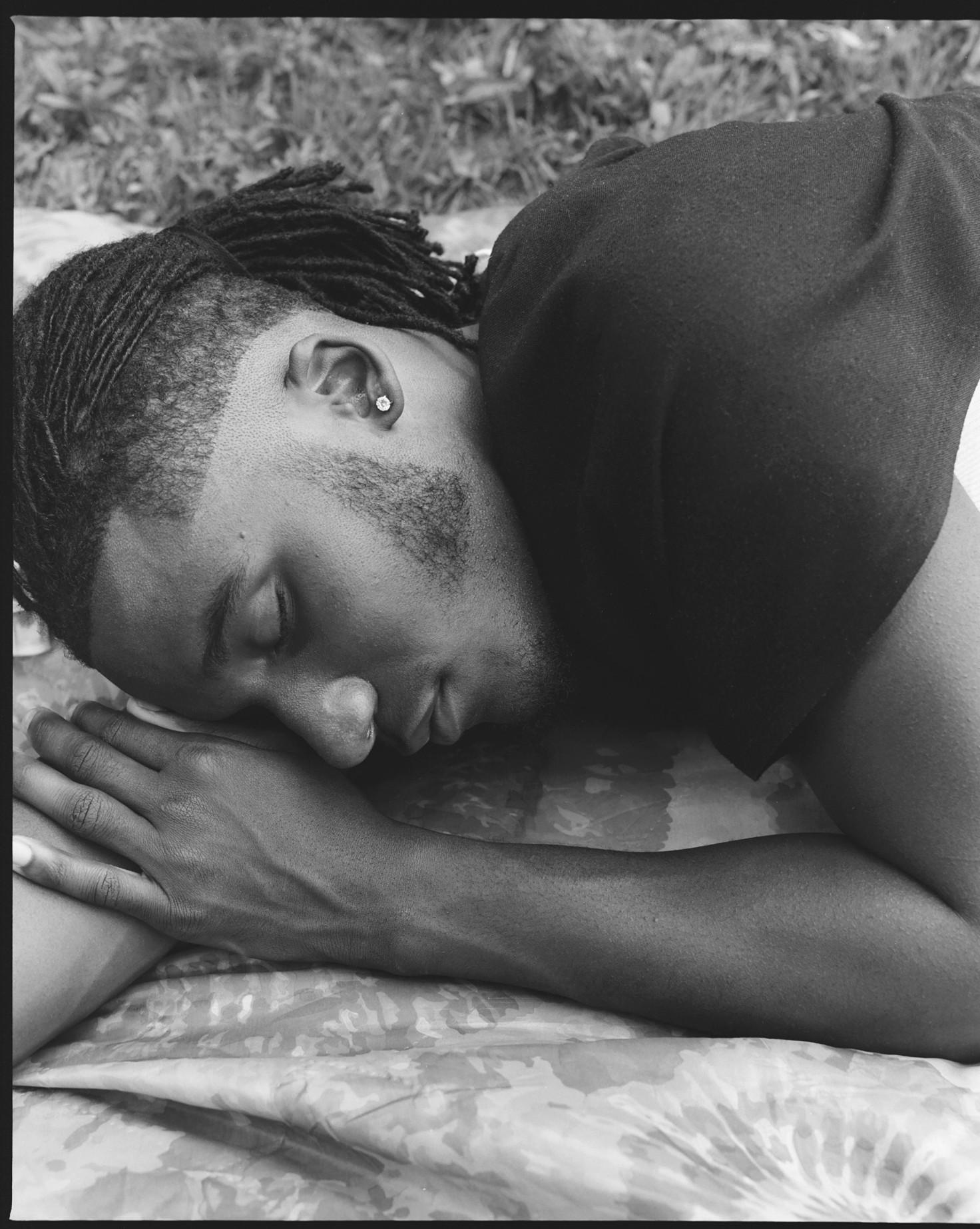 A Black person lays on a blanket on the ground. Their eyes closed and one arm resting on the other.