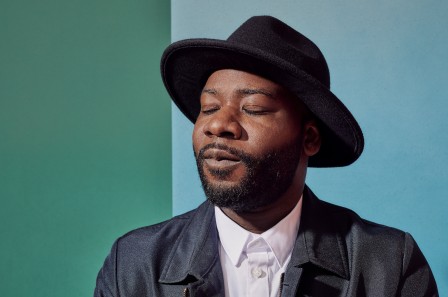 Blitz Bazawule wears a black rimmed hat and a black leather jacket over a white shirt, his eyes are closed as if in meditation. Behind him is a wall which is teal and light blue.