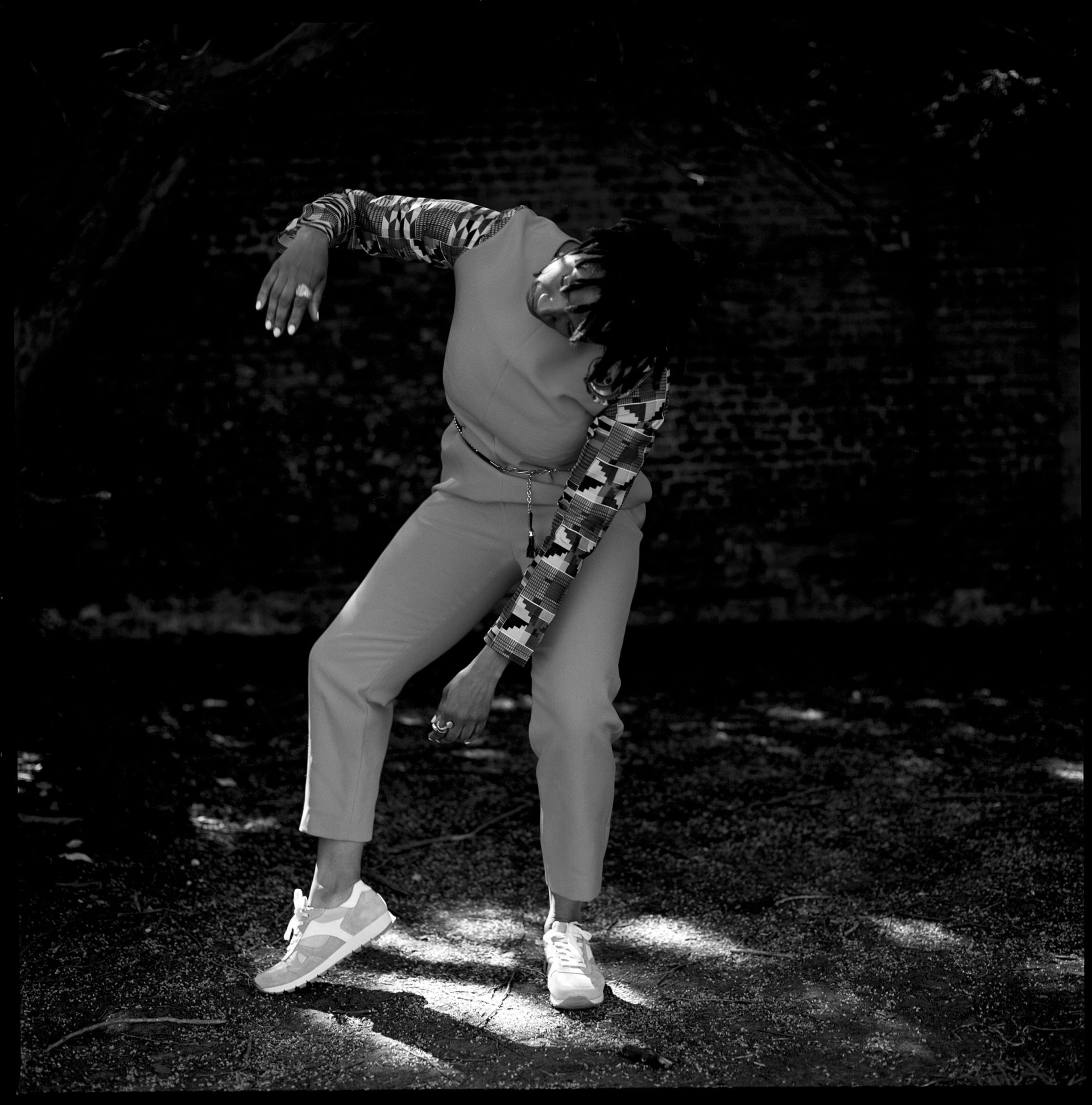 Black and white image of movement director Coral Messam. In it, she appears to be dancing, raising one arm up at a 90 degree angle and hanging her head. One foot is pointed out and she stands in front of a brick wall with vegetation surrounding her.