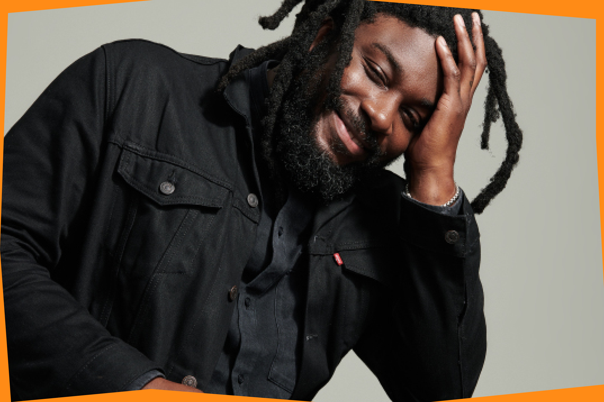 A photo of Jason Reynolds shows him wearing all black and smiling brightly towards the camera while resting his head in his left hand.