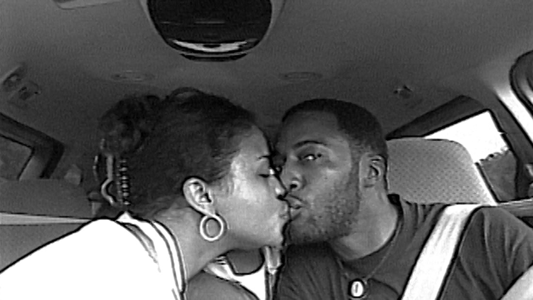 Still from "Time" is a black and white photo of a couple kissing in a car. As they turn towards each other, one has their eyes closed while the other's eyes are open and looking at the camera.