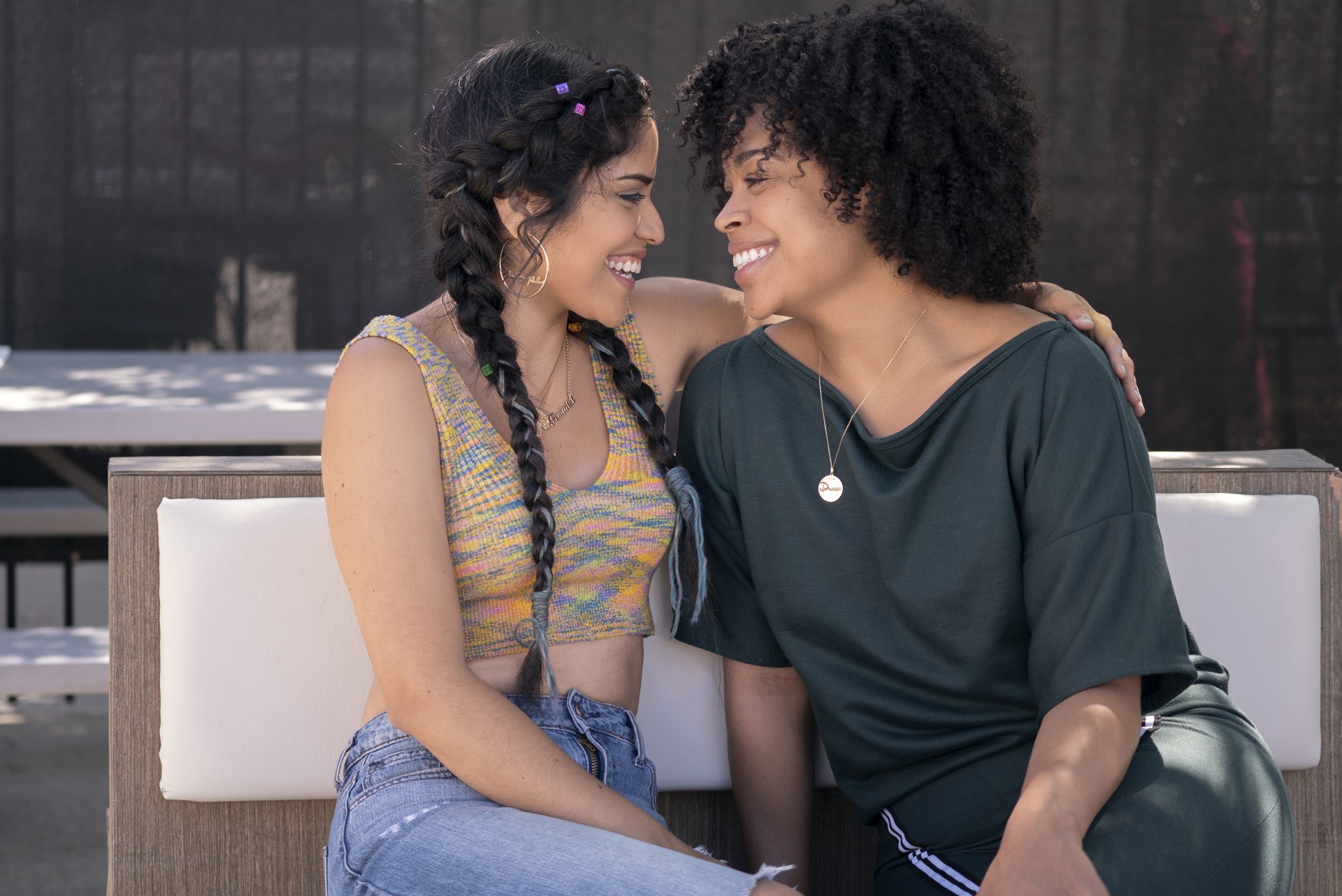 Still from the series "Gentefied" shows two people sitting on a bench. One wraps their arm around the other as they both lean in and smile at each other.