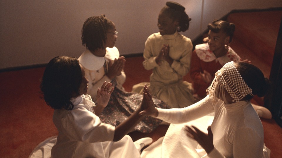 Five Black people sit in a circle playing a hand clapping game. Four of them are little girls, and one is an adult whose face is masked. They all wear white dresses.