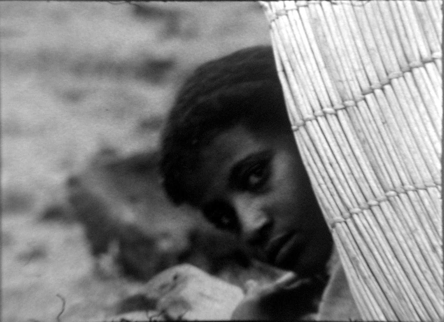 A Black person peers around a wicker object. In black and white.