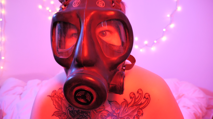 A person wearing a black gas mask looks into the camera. They do not appear to be wearing a shirt and a large floral tattoo can be seen on their chest.