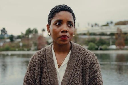 Janicza Bravo photo, wearing a cardigan sweater, standing in front of a body of water, looking off camera to the right.