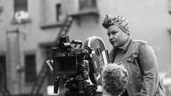 Image of a person standing in front of a large film camera on a film set. They wear a jean jacket and headscarf as they look at the camera.