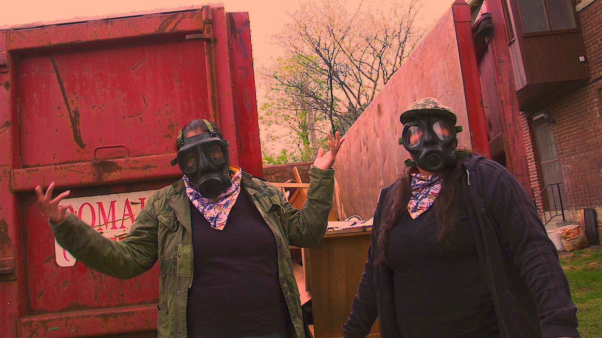 Image Description: Two women stand outside of a brick building. They are both wearing jackets, black T-shirts, colorful scarves around their necks, and gas masks that obscure most of their faces.