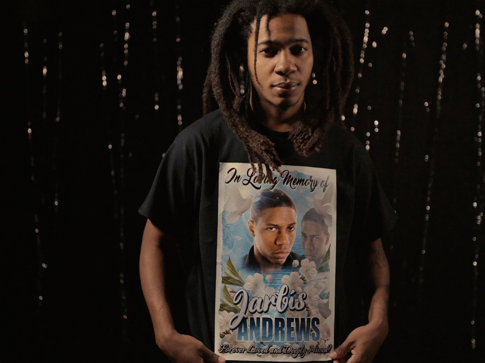 Still from the film "T" shows a young black man with dreadlocks standing in front of a twinkling backdrop. He wears a shirt from a memorial of a loved one with a large photo that says "In Loving Memory of Jarbis Andrews."
