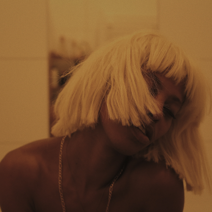 A still image from Rikki Wright's "A Song About Love" shows a Black person in a blonde wig, seemingly in a state of ecstasy or mid-dance, their head tilted to the side and hair obscuring much of their face.